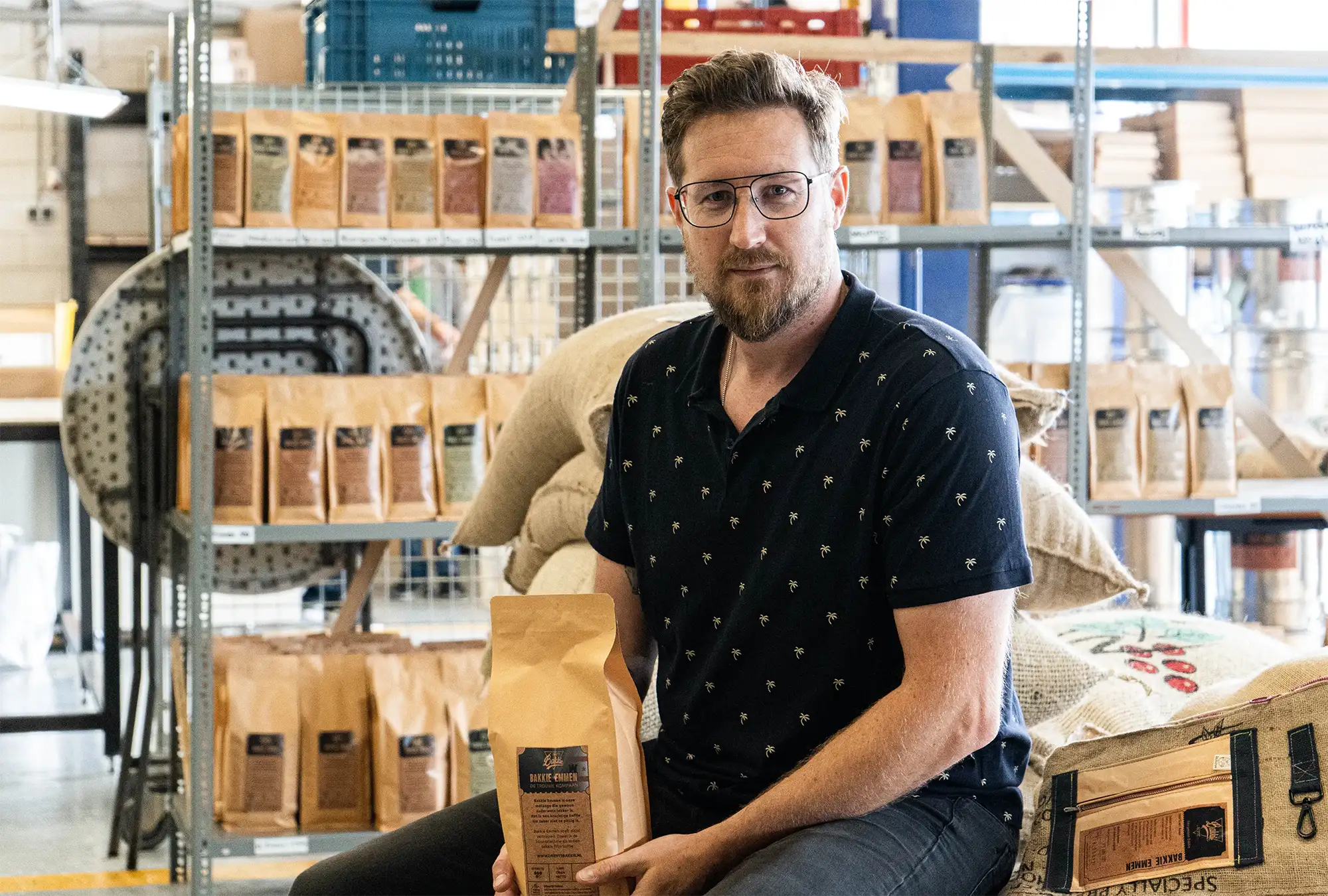 Man met bril poseert voor zakken Drentse Koffie terwijl hij een zak koffie vasthoudt.